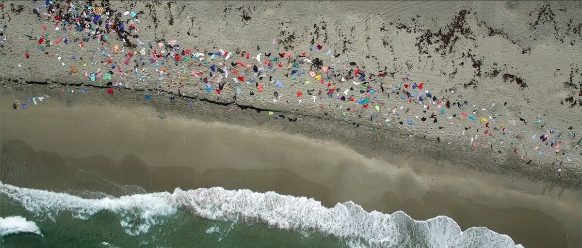 Image depicting colorful clothing littered like confetti on a sandy shoreline. Still from Rest Ashore video projection. Image courtesy of Juana Valdes.